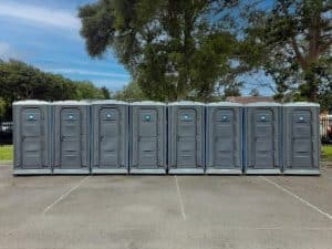 A row of grey portable toilets lined up in an outdoor parking area, surrounded by trees and greenery in the background.